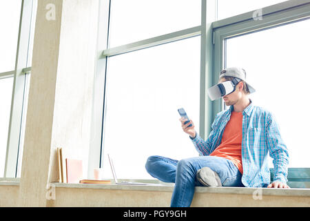Serious student boy in VR headset sitting on window sill and using gadget to operate virtual reality simulator in university corridor Stock Photo