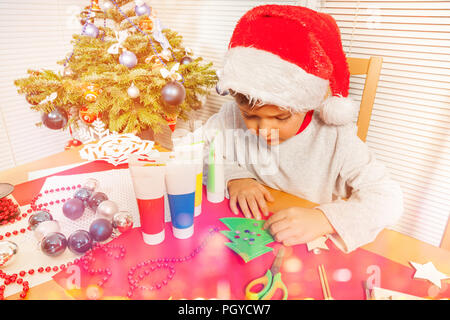 Portrait of little boy in Santa's hat decorating paper Christmas tree, sitting at the desk in the kindergarten Stock Photo