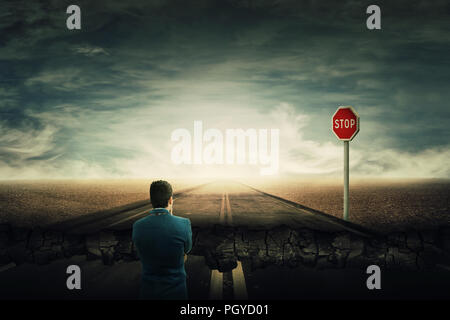 Surreal view as a man stand on a crushing asphalt road and a STOP red sign in front. Deep hole in the ground, chasm as broken way for businessman. Car Stock Photo