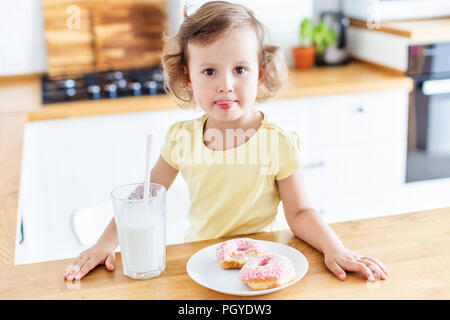 Little girl eating donuts and drinking milk on the white kitchen at home. Child is having fun with donuts. Tasty food for kids. Stock Photo