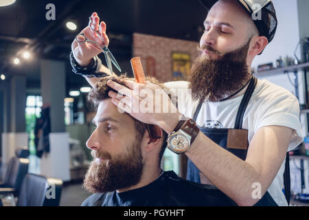 Dedicated male hairstylist using scissors and plastic comb while giving a cool haircut to a redhead bearded young man in a trendy beauty salon Stock Photo