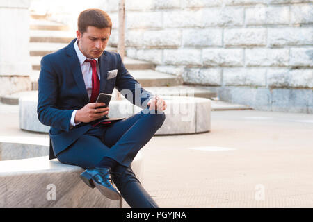 Young handsome businessman in a suit and tie consults the smartphone sitting on a marble bench in the park Stock Photo