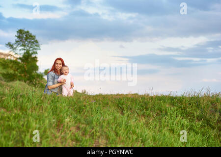 Young mother hugging cute little daughter sitting on green grass in field. Mom and child feeling happy, smiling, looking at camera, spending family ti Stock Photo