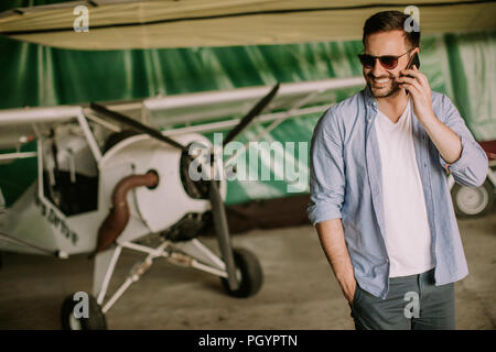 Young handsome young pilot checking airplane in the hangar and using mobile phone Stock Photo
