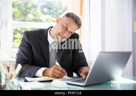 Businessman Talking On Cellphone And Using Laptop While Writing Schedule In Diary Stock Photo