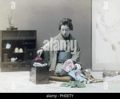 [ 1890s Japan - Japanese Woman in Kimono Sewing ] —   A young Japanese woman in kimono and traditional headwear is sewing clothes for dolls while sitting on a zabuton cushion. Two dolls are leaning against her knees. A sewing kit with a pin cushion stands on the floor in front of her.  19th century vintage albumen photograph. Stock Photo
