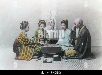 [ 1890s Japan - Playing Go ] —   Three young women in kimono and traditional hairstyles and an elderly man in a haori coat are seated around a board for the game of go.  This studio is arranged as a zashiki (guest room), with an ikebana flower arrangement and scroll in the back. A small tray with a teapot and two cups can be seen at the front.  19th century vintage albumen photograph. Stock Photo