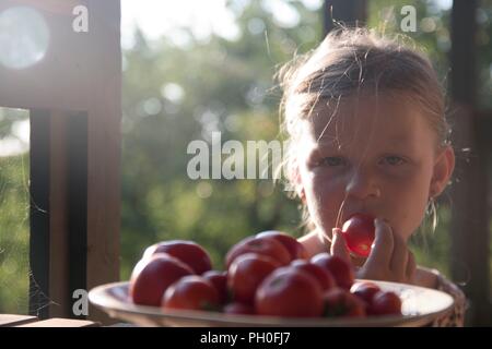 The little girl is sitting at a table on a verandah and eatting fresh tomatoes Stock Photo