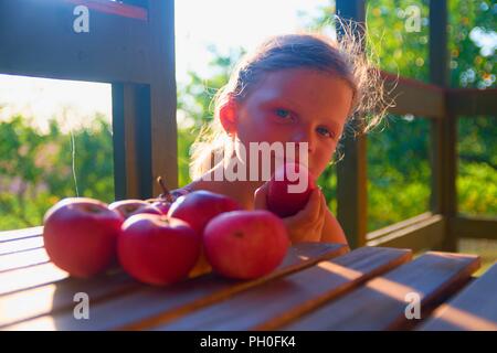 Little girl is sitting on porch in summer. Small girl is eating apples. Apples on table. Dreamy and romantic image. Summer and happy childhood concept Stock Photo