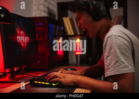 Portrait of asian tense angry gamer boy losing while playing video games on computer in dark room wearing headphones and using backlit colorful keyboa Stock Photo