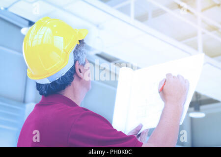 asian engineer in yellow hard safety helmet is checking the structure of building and factory blueprint on the factory construction site with sunbeam. Stock Photo