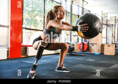 Portrait of european invalid woman with prosthesis in tracksuit doing sit-ups with fitness ball in gym Stock Photo