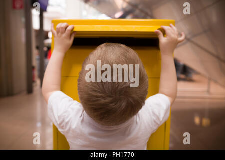 A little blond boy in a white t-shirt puts a letter into a yellow postbox. Sending post to a friend at the post office. Stock Photo