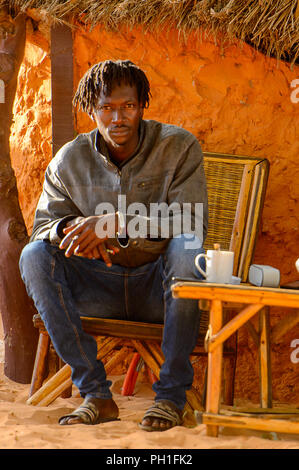 LAMPOUL DESERT, SENEGAL - APR 23, 2017: Unidentified Senegalese man with braids sits near the shack in a Lampoul Desert, beautiful landscape place Stock Photo