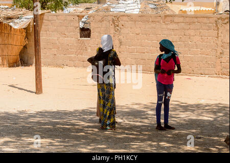 ROAD TO LAMPOUL, SENEGAL - APR 23, 2017: Unidentified Senegalese two girls with backpacks stand in the shadow from the tree. Still many people in Sene Stock Photo