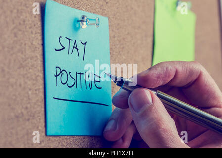 Retro instagram style image of a male hand writing Stay positive on blue post it paper pinned on cork bulletin board. Stock Photo