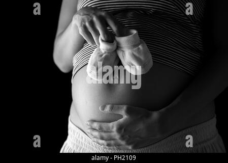 Pregnant woman holding a pair of cute baby booties above her swollen abdomen as she awaits the birth of her unborn child, shadowy black and white clos Stock Photo