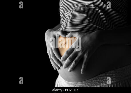Pregnant woman making a heart gesture on her belly with her fingers - selective colour of the heart shape in a greyscale image conceptual of love of h Stock Photo