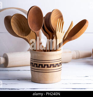 various wooden objects in a wooden jar, spoons, forks, rolling pin, plates and a sieve on a white wooden table Stock Photo