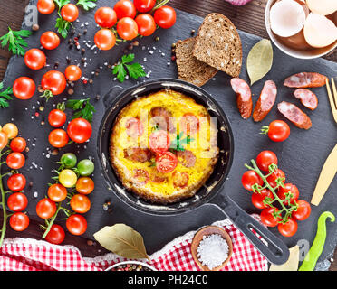 fried omelette from chicken eggs with red cherry tomatoes and smoked sausage in a round black frying pan on a table in the middle of vegetables, top v Stock Photo