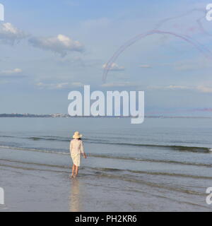 Dorset, UK, 30th August 2018. The Red Arrows aerobatic display team perform against blue skies on the first day of the Bournemouth Air Show. Woman standing in the sea and watching from Shell Bay, Studland. Stock Photo