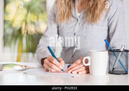 Business woman writing in document with pen at desk in modern office. Girl wears elegant shirt on strips. Big green blurred windows in background