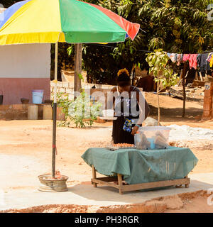ROAD TO BISSAU, GUINEA B. - MAY 1, 2017: Unidentified local woman stands near the table and umbrella in a village in Guinea Bissau. Still many people  Stock Photo