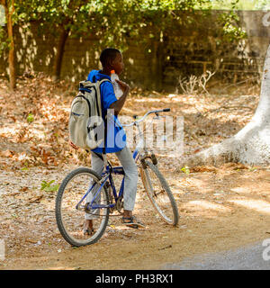 ROAD TO BISSAU, GUINEA B. - MAY 1, 2017: Unidentified local  boy with backpack holds a bicycle in a village in Guinea Bissau. Still many people in the Stock Photo
