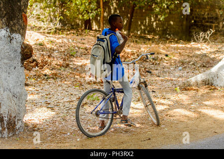 ROAD TO BISSAU, GUINEA B. - MAY 1, 2017: Unidentified local  boy with backpack holds a bicycle in a village in Guinea Bissau. Still many people in the Stock Photo