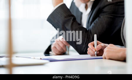 Business partners, sitting in an office checking a document or contract while signing it, cropped side view. Stock Photo