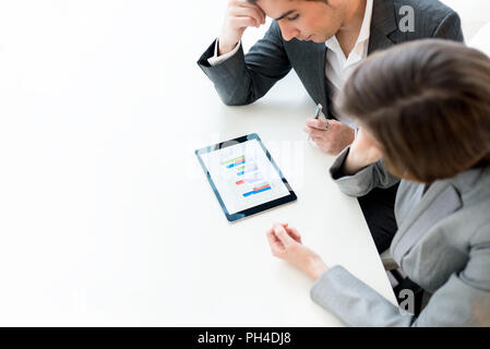 Overhead view of two business colleagues, a man and woman, analysing a statistical business graph on a tablet computer as they sit side by side at the Stock Photo