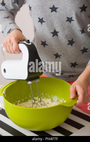 Young woman in long sleeve shirt with stars mixing and tasting dough for homemade muffins. Cooking and baking at home. Bright kitchenware and white mi Stock Photo