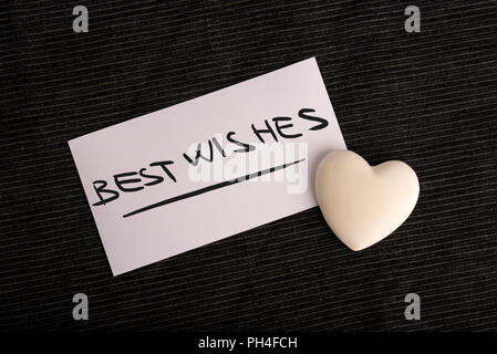Best wishes for a loved one with a hand-written plain white card saying - Best Wishes - with a cream colored heart on a black background, overhead vie Stock Photo