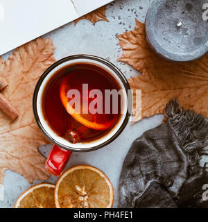 Autumn season tea with cinnamon and orange slice surrounded by black aromatic candles, warm scarf, maple leaves and open notebook on a concrete grey b Stock Photo