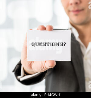 Businessman holding up a small white card saying Questions in a conceptual image of customer support and client services, close up of his hand. Stock Photo