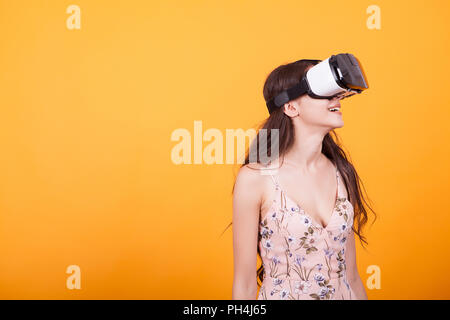 Happy girl getting experience using VR headset glasses of virtual reality in studio over yellow background. Stock Photo