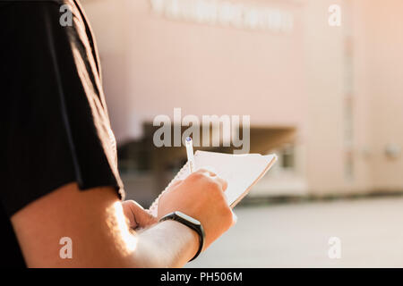 Man taking notes outside an educational facility. Young male person's hands holding a notepad and pen and writing information outdoors Stock Photo