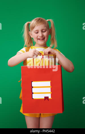 smiling pupil with backpack looking inside shopping bag with stationery against green background Stock Photo