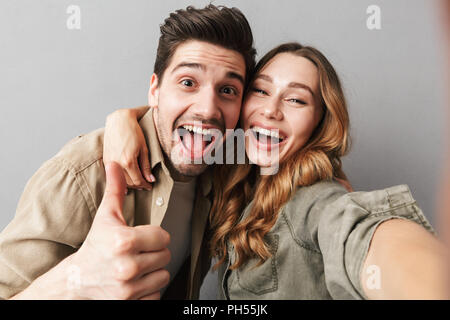 Portrait of an excited young couple hugging while taking a selfie and showing thumbs up isolated over gray background Stock Photo