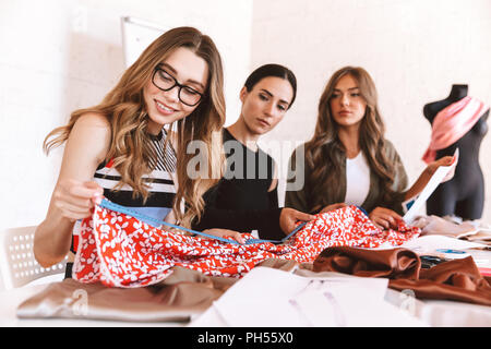 Three attractive young women clothes designers working together at the atelier, sitting at the table Stock Photo