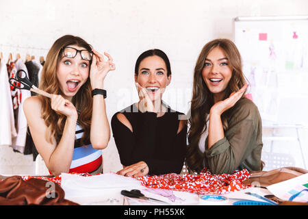Three joyful young women clothes designers working together at the atelier, sitting at the table Stock Photo