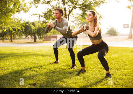 Image of young caucasian sporty man and woman 20s in tracksuits doing workout and squatting together in green park during sunny summer day Stock Photo