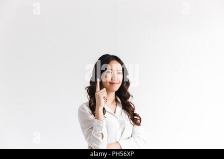 Young asian woman with long dark hair looking at camera with beautiful smile and pointing finger upward at copyspace text or product isolated over whi Stock Photo