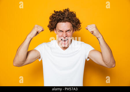 Photo of strong curly man 20s in casual t-shirt screaming and showing biceps at camera isolated over yellow background Stock Photo