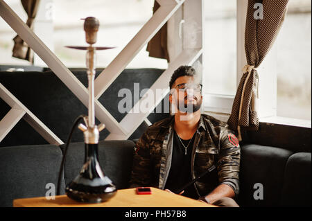 Stylish beard arabian man in glasses and military jacket smoking hookah at street bar. Arab model having rest. Stock Photo