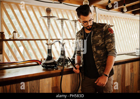 Stylish beard arabian man in glasses and military jacket smoking hookah at street bar. Arab model having rest. Stock Photo