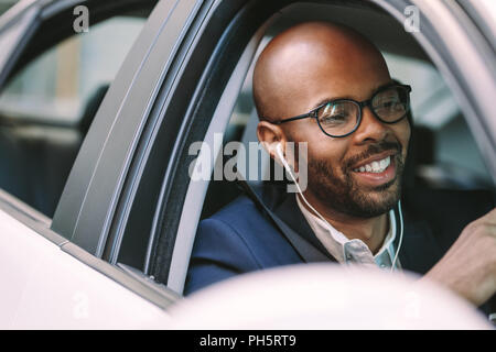 Young bald man smiling inside the car while driving. African man in suit and wearing earphones enjoying driving a car. Stock Photo