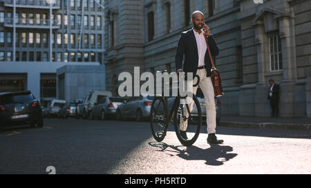 Young businessman walking outdoors with a bicycle and using mobile phone. African man going to work on bike talking over cell phone on city street. Stock Photo