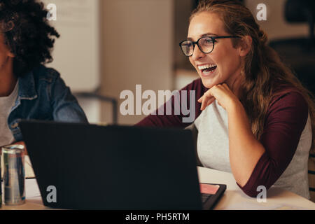 Woman sitting at table with laptop looking away and laughing wit colleague sitting by. Female software engineer laughing in office. Stock Photo