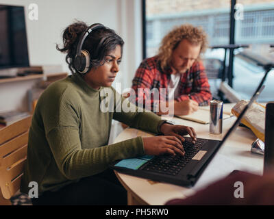 Computer programmers wearing headphones coding on laptop with colleagues working around at startup office. Young professionals working at a tech start Stock Photo